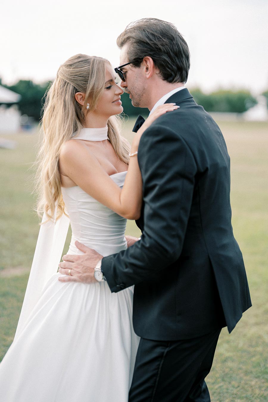 Elegant couple sharing a romantic moment outdoors, woman in a white strapless dress and man in a black suit embracing on a grassy field