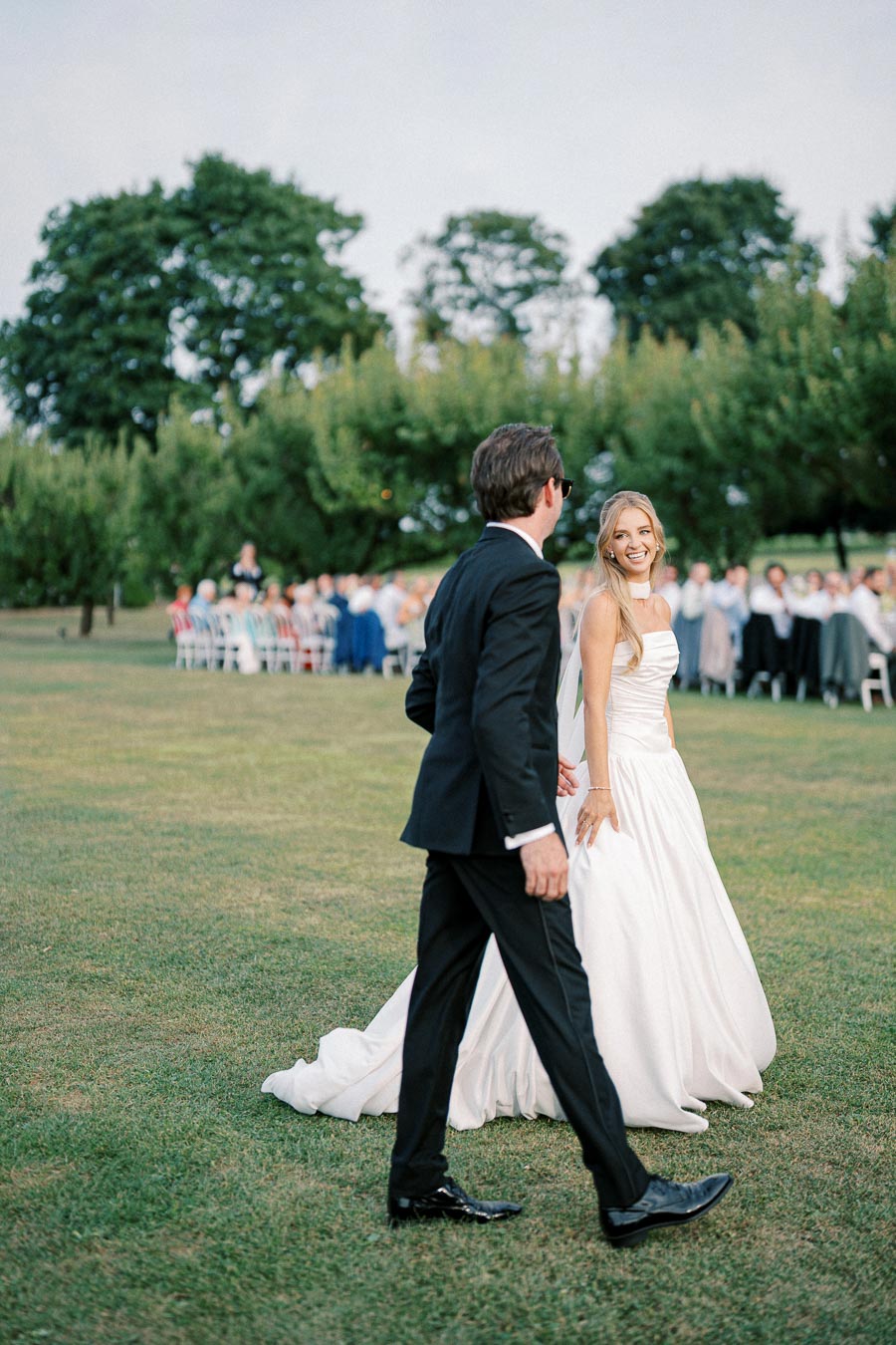 A bride in a white wedding dress and a groom in a black suit walk across a lush green lawn during an outdoor wedding ceremony, with guests seated in the background under a canopy of trees.