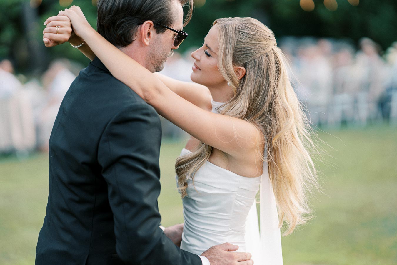 Elegant couple dancing closely at an outdoor wedding, the woman in a white gown and the man in a black suit, surrounded by greenery.
