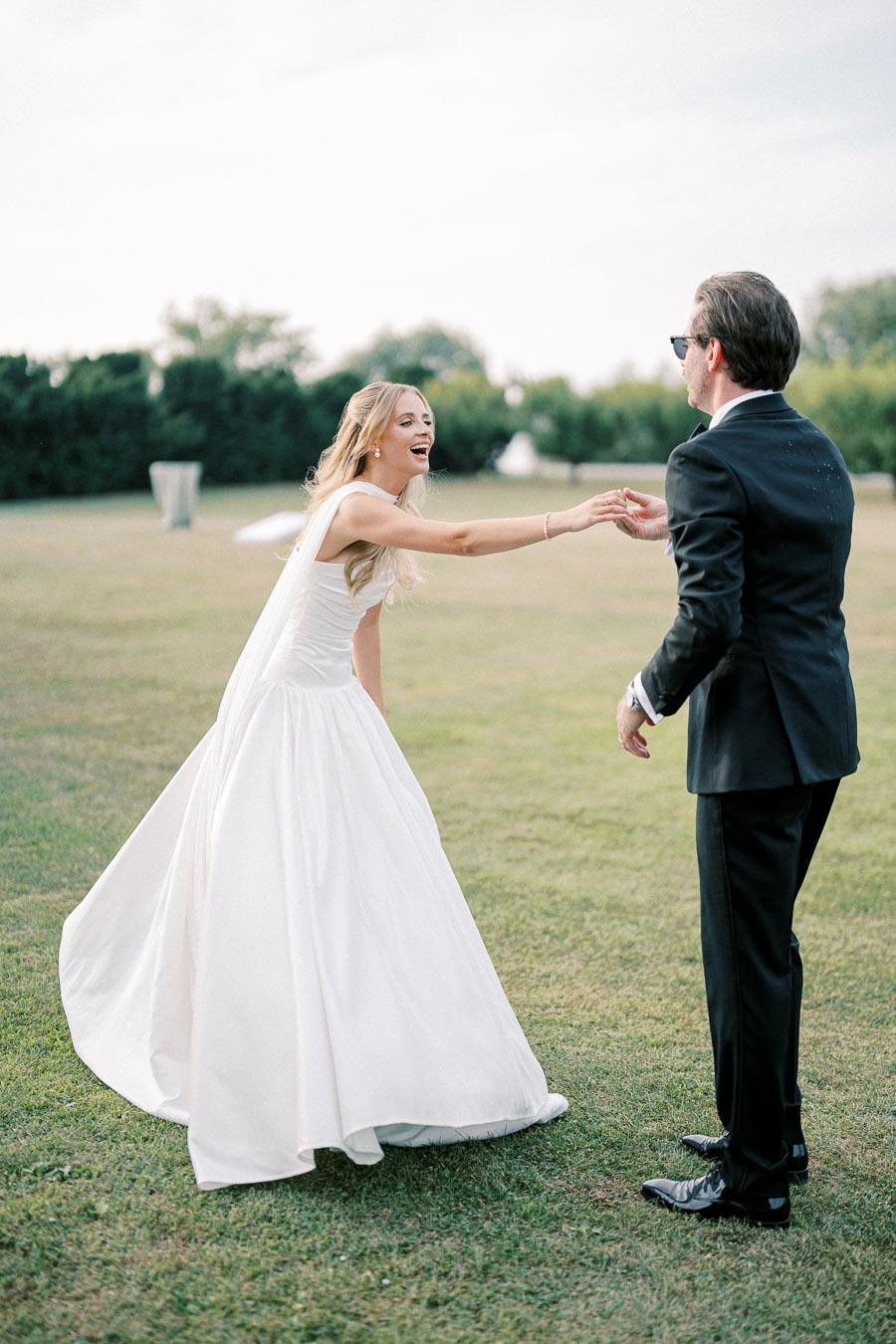 A joyful bride in a flowing white dress dances with her partner in a suit on a grassy outdoor venue.