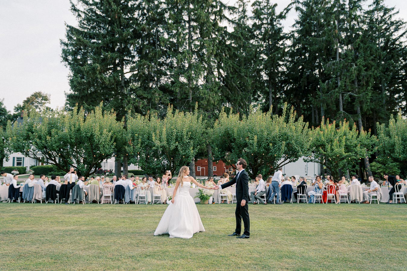 A bride and groom dancing outdoors at a wedding reception, surrounded by seated guests under large, lush trees on a bright, clear day.