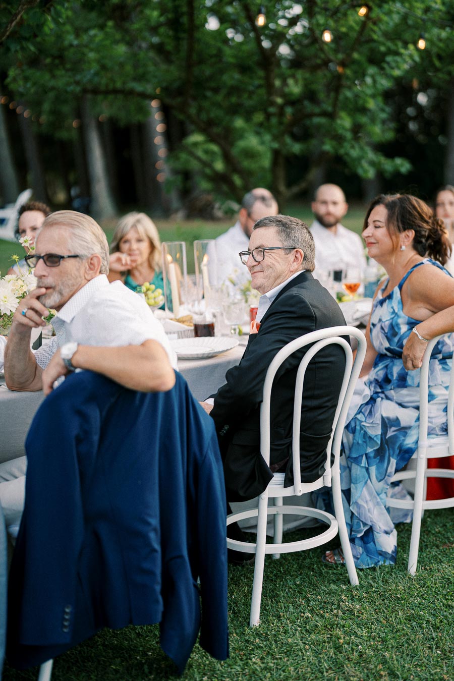 Group of people sitting at an outdoor wedding reception, smiling and enjoying the event under string lights with a green, wooded background.