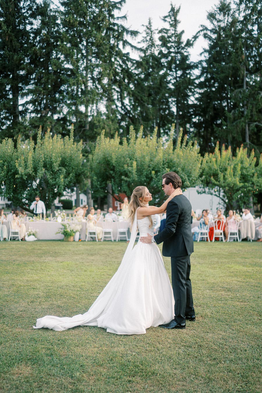 A bride in a flowing white gown and groom in a black suit share a romantic dance on a lush green lawn, surrounded by guests seated at tables, with a backdrop of tall trees under a clear sky.