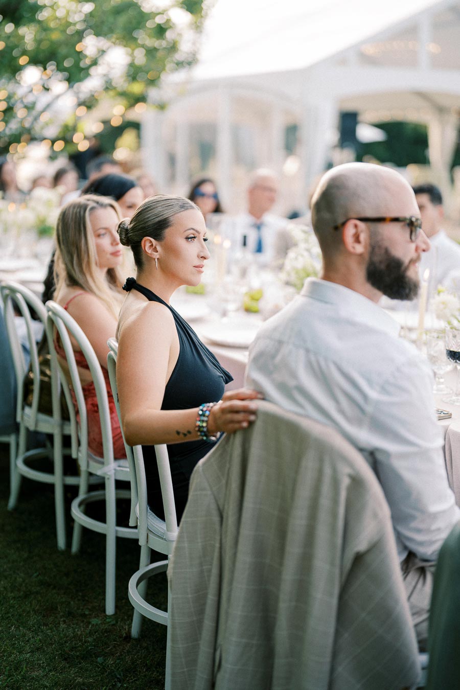 Group of elegantly dressed people seated at an outdoor wedding reception, attentively listening during a speech.