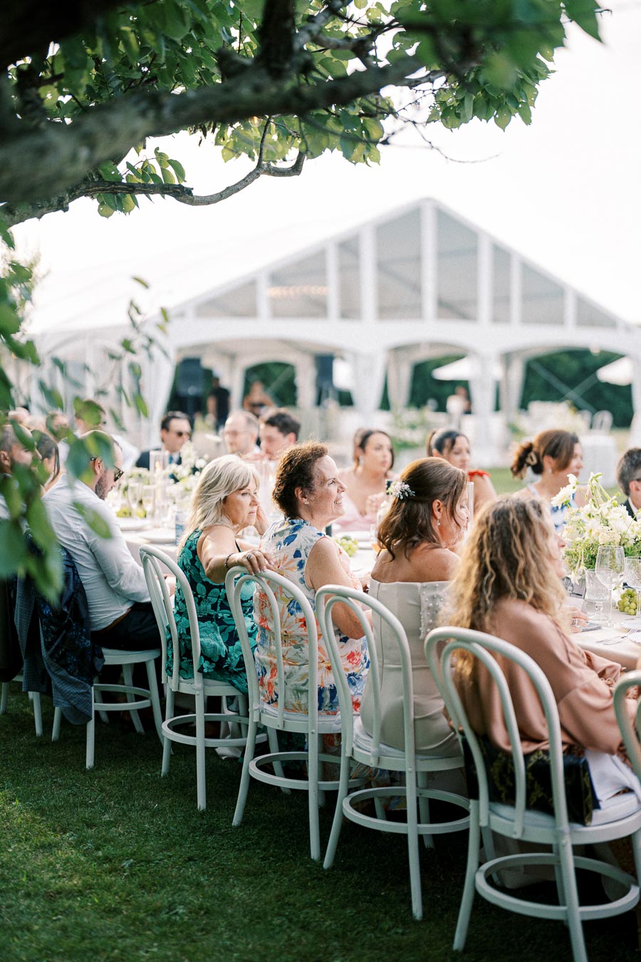 Outdoor wedding reception with elegantly dressed guests seated around a table under a white tent on a sunny day.