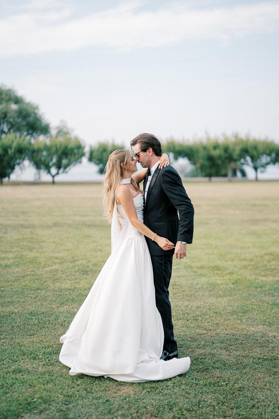 A bride in a white gown and a groom in a black suit embrace on a grassy field with trees in the background, under a partly cloudy sky.
