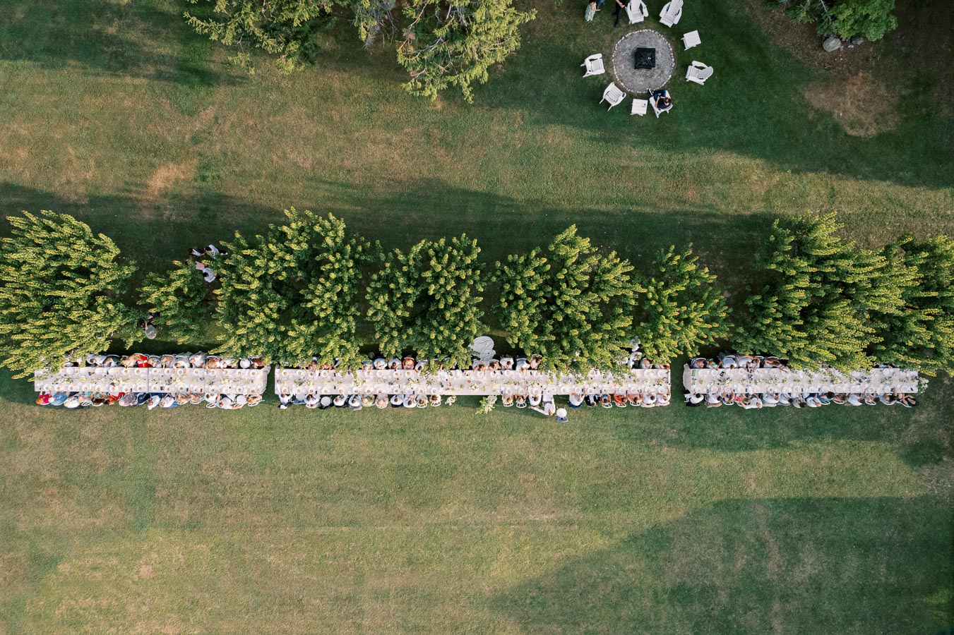 Aerial view of a long outdoor dining table set in a green garden, surrounded by lush trees, with guests enjoying a communal meal.