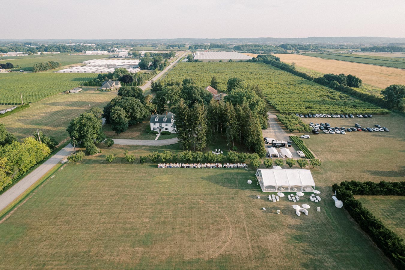 Aerial view of a large rural property with a country house surrounded by greenery and fields. A wedding setup is visible with white tents and seating arrangements on the lawn. Several cars are parked near the fields, suggesting an event or gathering in progress.