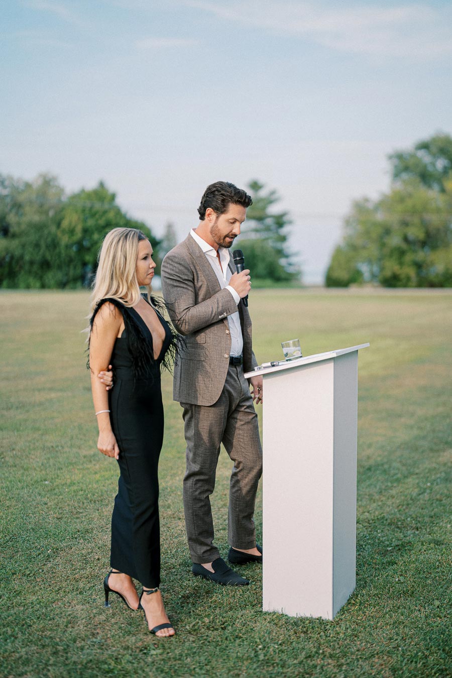 Man in a suit speaking at a podium outdoors with a woman in a black dress standing beside him, set against a background of green grass and trees.