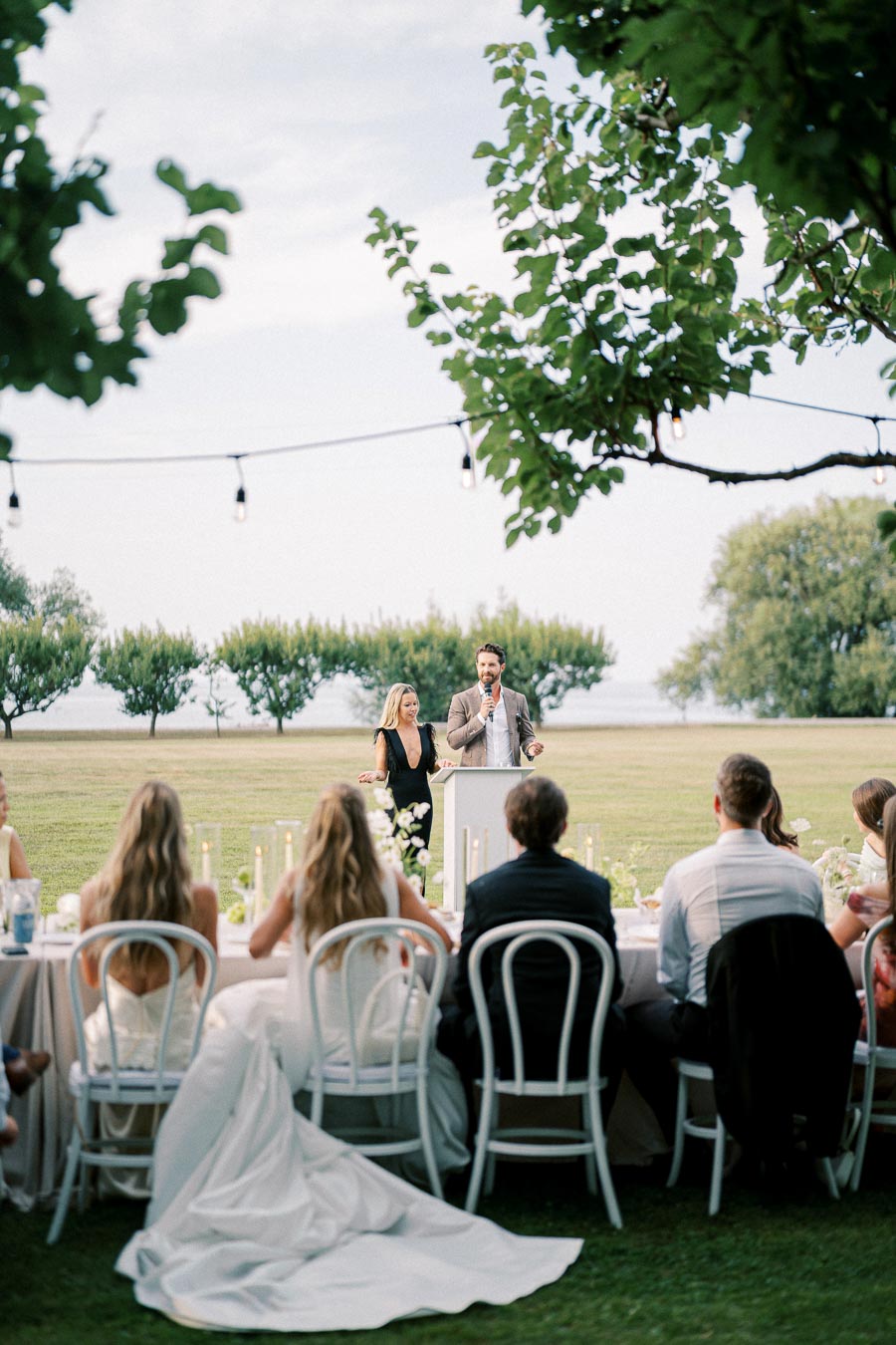 Outdoor wedding reception scene with a group of guests seated at an elegantly decorated table, listening to a speech by a couple standing at a podium, surrounded by lush greenery and string lights.
