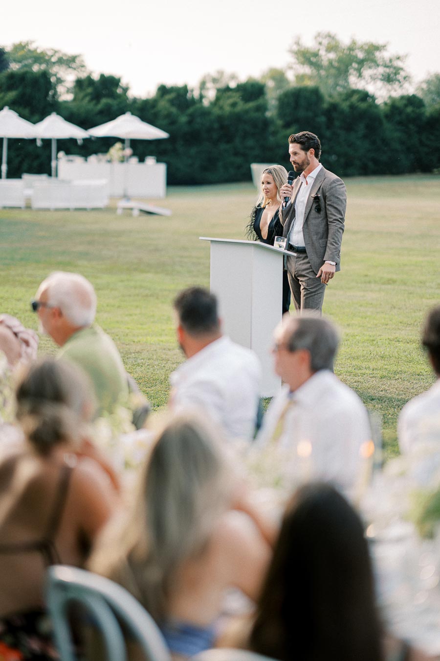 Two people speaking at a podium during an outdoor event with attendees seated at tables, green lawn, and white umbrellas in the background.