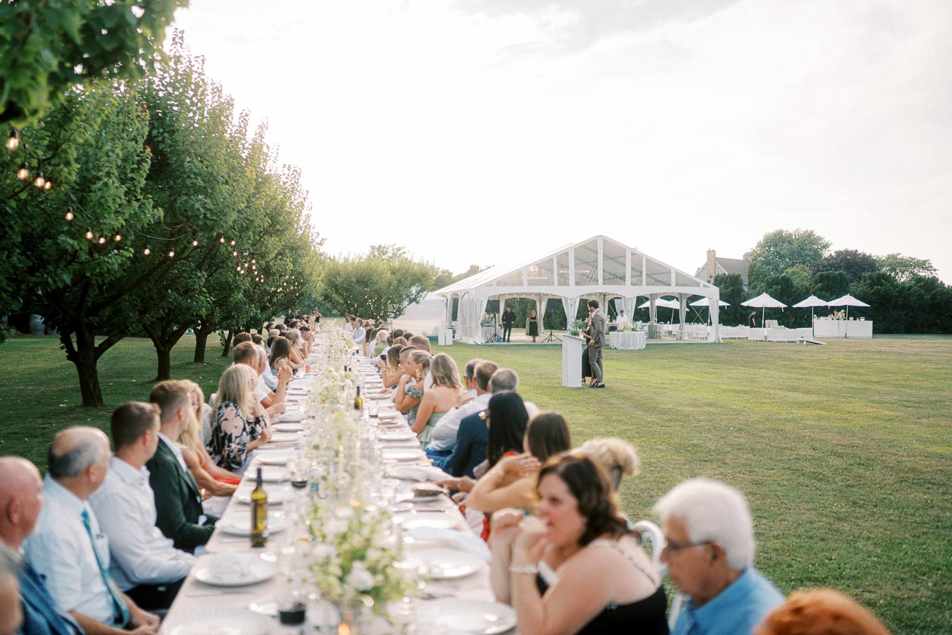 Outdoor wedding reception with a long table of seated guests in a garden setting, featuring a large white tent and lush greenery, creating a scenic and elegant atmosphere.