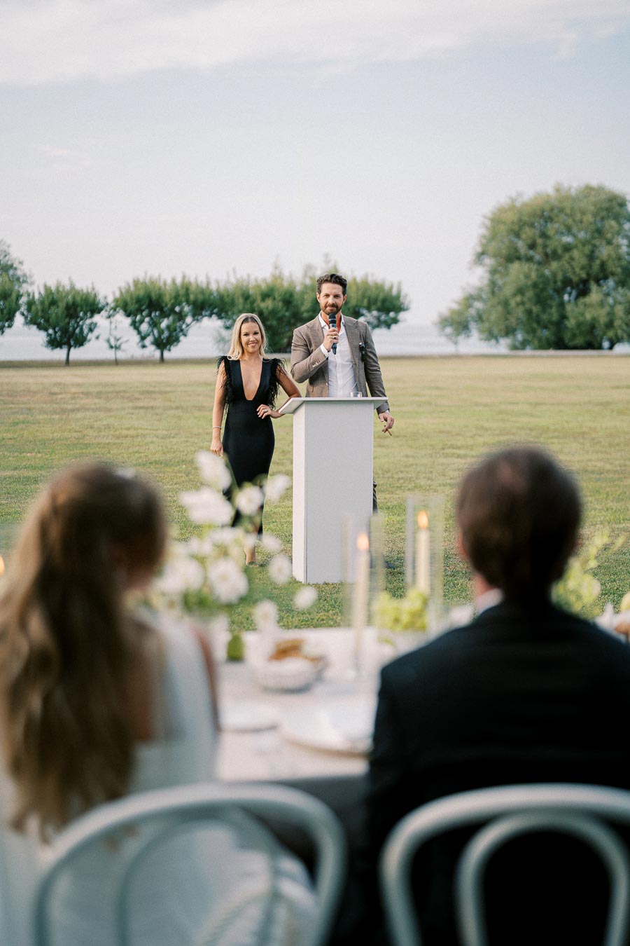 A couple delivering a speech at an outdoor wedding reception, standing at a podium on a grassy field with guests seated at tables in the foreground, decorated with white flowers and candles.