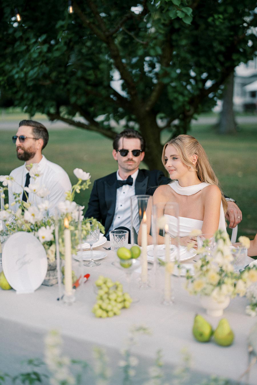 Elegant outdoor wedding reception with guests seated at a beautifully decorated table featuring white flowers, candles, and fresh fruits, set against a lush green garden backdrop.