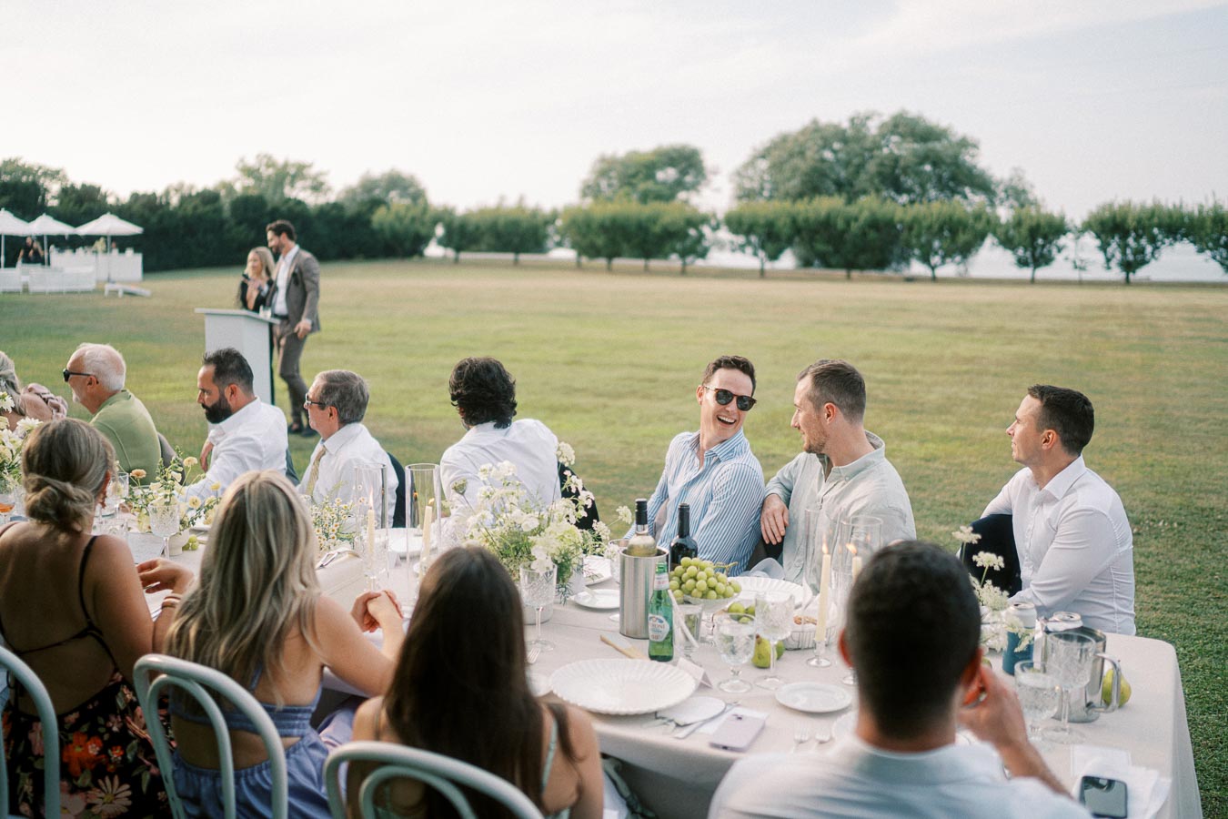 Outdoor wedding reception with guests seated at a table decorated with flowers and wine, listening to a speech in a scenic garden setting.