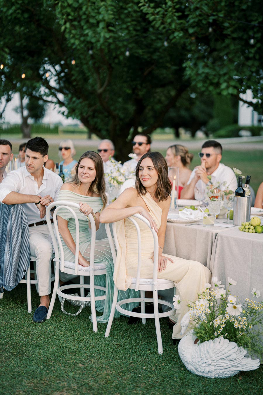 A group of elegantly dressed people seated at an outdoor wedding reception, surrounded by greenery and a beautifully set table adorned with flowers and candles.