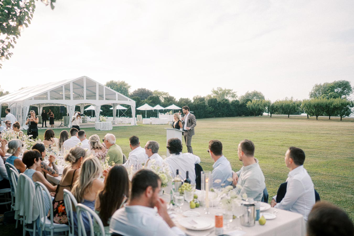 Outdoor wedding reception with guests seated at a long table, listening to a speech by a couple at a podium, surrounded by a scenic garden and white tent pavilion.