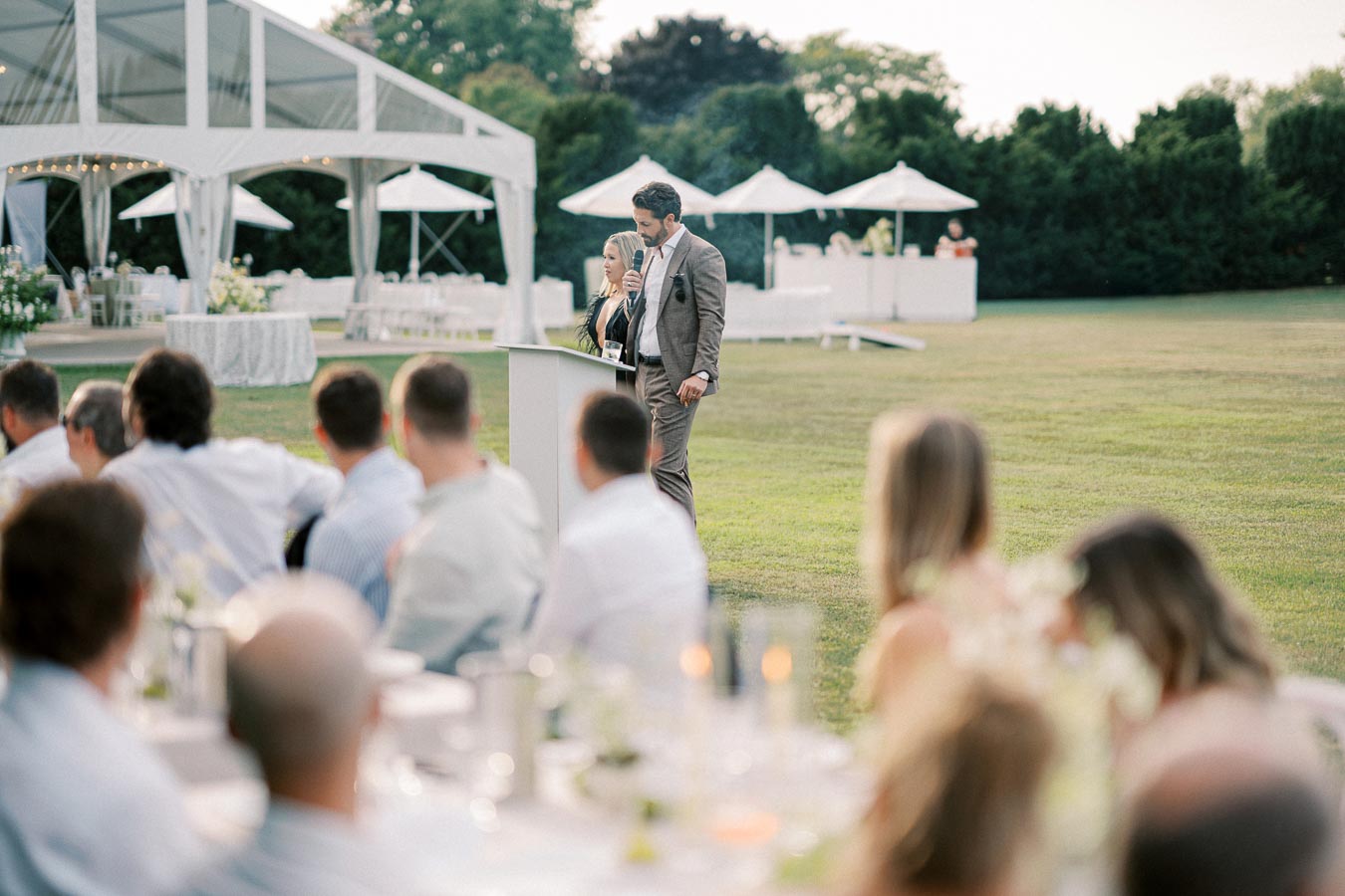 Outdoor wedding reception with a speaker at a podium addressing seated guests, elegant white canopy and umbrellas in the background, on a sunny day.