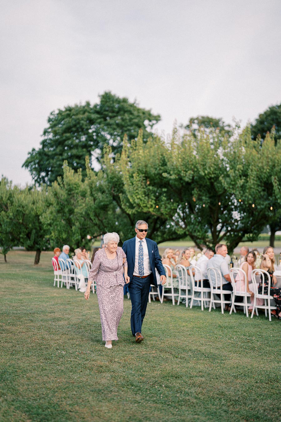 Elderly woman and man walking together at an outdoor wedding ceremony, surrounded by seated guests and lush green trees.