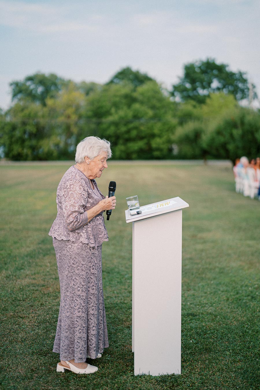 Elderly woman speaking at an outdoor event podium, wearing a lavender lace dress, with greenery in the background.