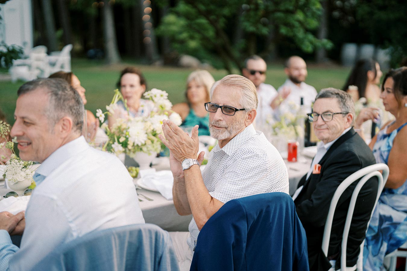 Group of people sitting at a beautifully set outdoor event, with a man in the foreground clapping and others engaged in conversation around a table adorned with floral arrangements.