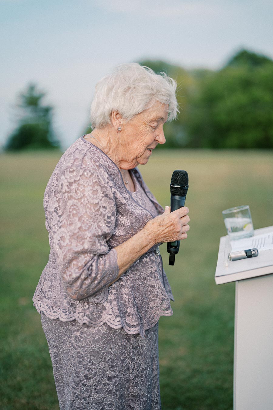 Elderly woman in a lavender lace dress giving a speech outdoors, holding a microphone near a podium with a drink and papers on it, with green grass and trees in the background.