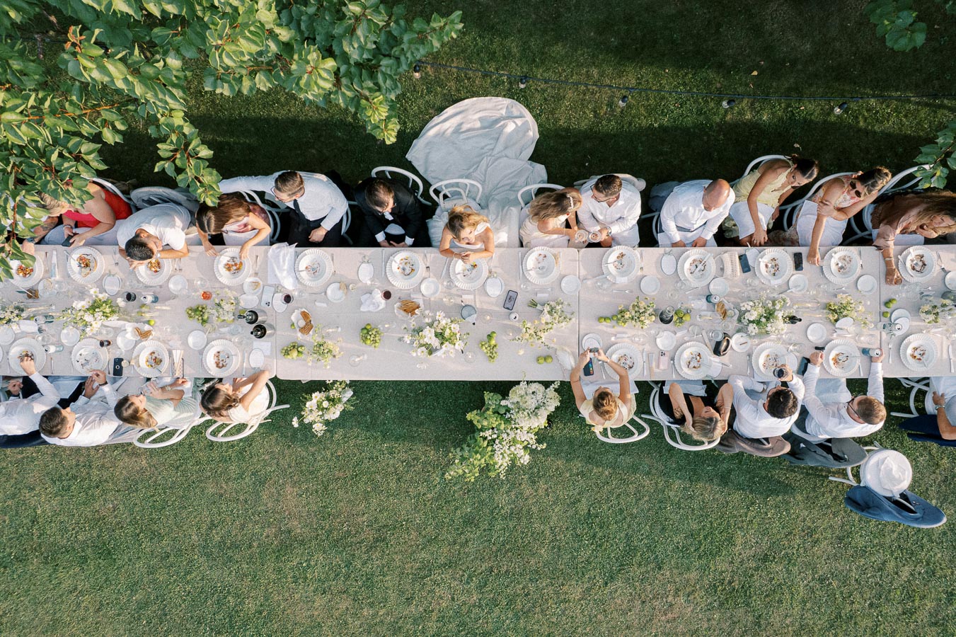 Aerial view of an elegant outdoor dining table set for a wedding reception, adorned with white floral arrangements, surrounded by guests in formal attire enjoying a meal on a grassy lawn.