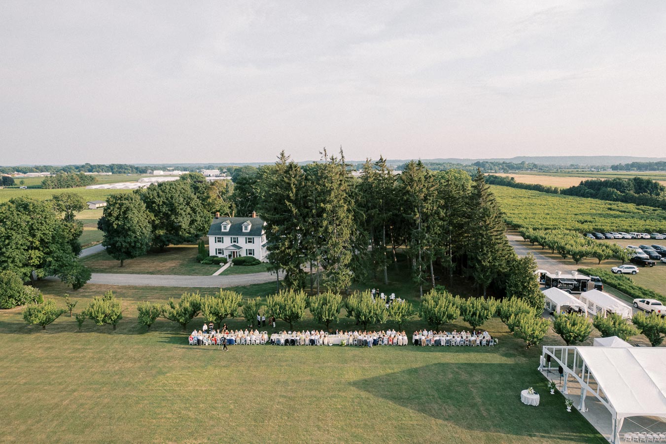 Aerial view of a countryside wedding reception with long banquet tables and guests seated on an expansive green lawn. A charming white house with a dark roof is nestled among tall trees, with sprawling fields in the background under a partly cloudy sky.
