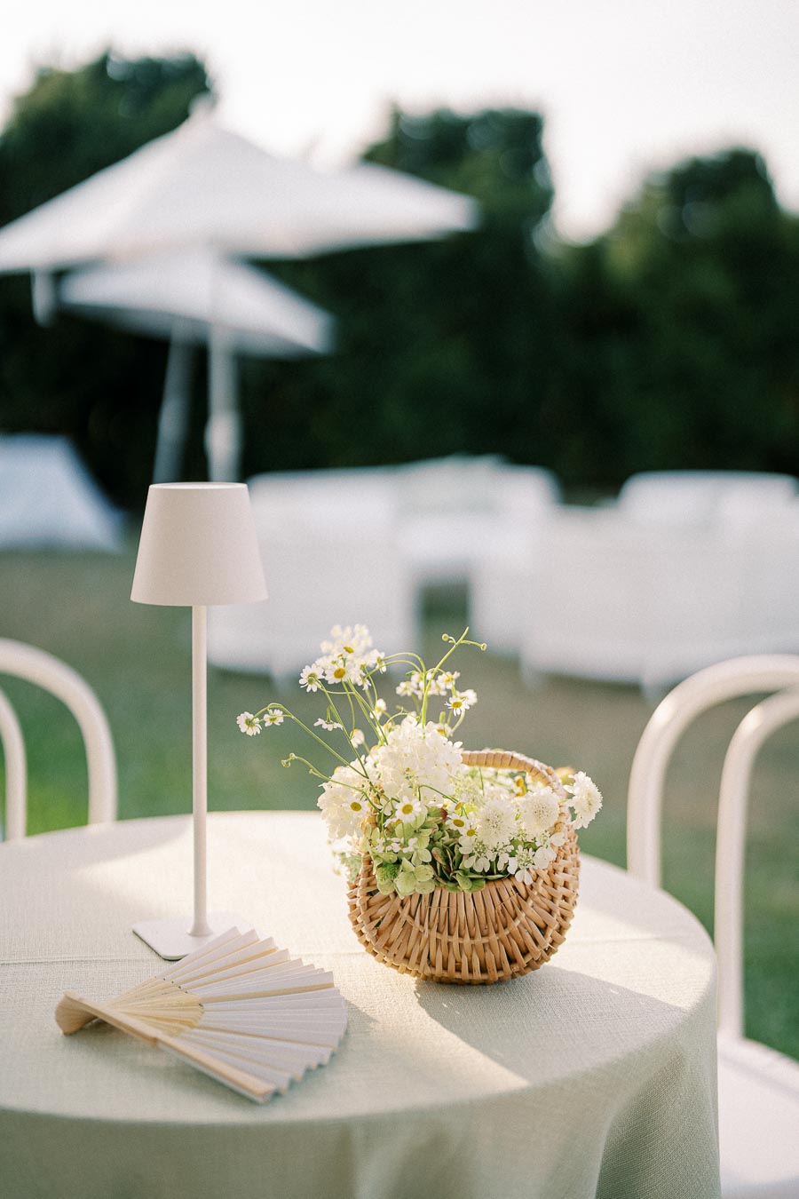 Outdoor dining setup with a round table featuring a wicker basket of white daisies, a small white lamp, and a paper fan, set against a blurred background of green trees and white umbrellas. Perfect for a garden party or elegant event.