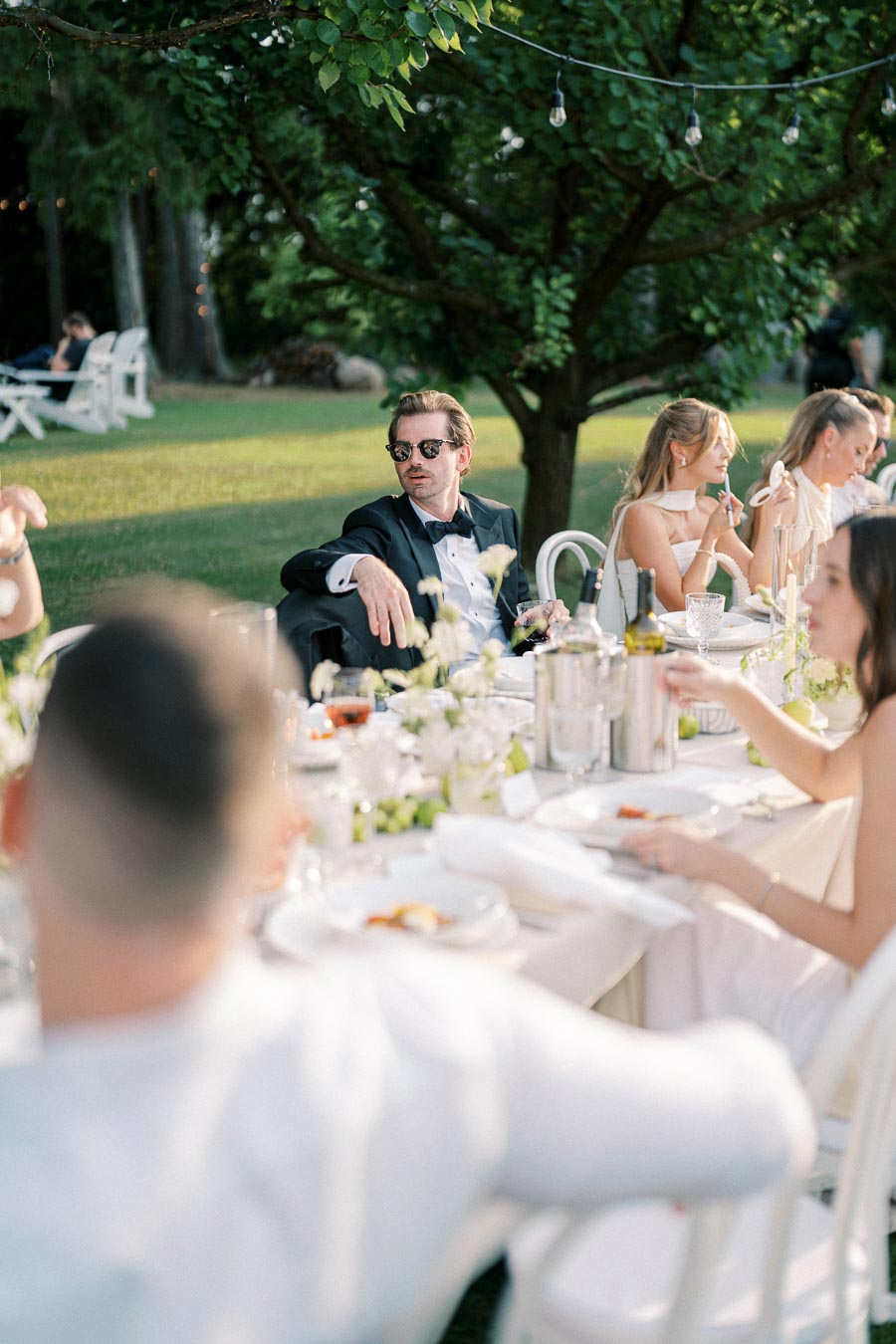 Elegant outdoor gathering with guests seated at a beautifully decorated table under lush green trees, featuring a man in a tuxedo and sunglasses engaging with others at the event.