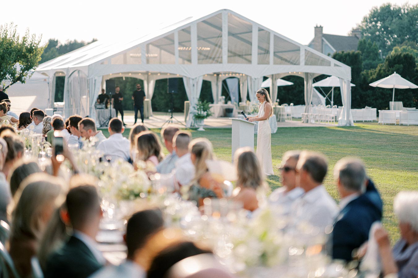 Outdoor wedding reception with a woman giving a speech under a large white tent, surrounded by seated guests at elegantly decorated tables.