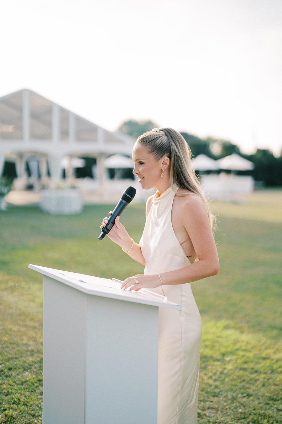 Woman in an elegant white dress delivers a speech outdoors at a podium with a microphone, set against a bright, airy outdoor event backdrop.