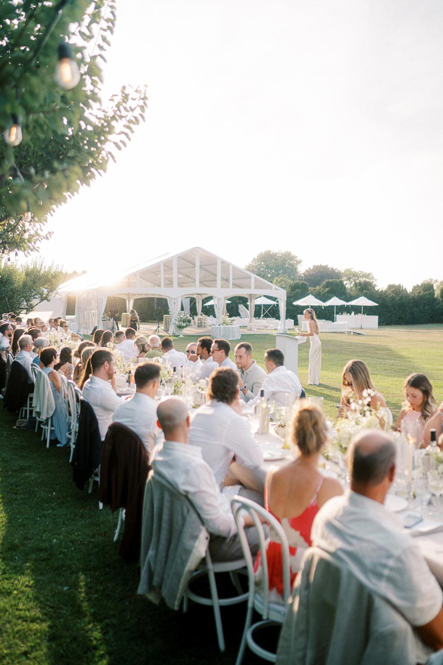 Outdoor wedding reception with guests seated at a long, elegantly decorated table under a clear sky, near a large white canopy in a lush green field.