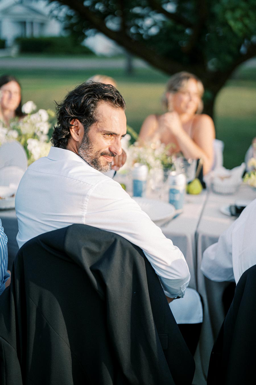 A man in a white shirt smiling while seated at an outdoor event table with blurred guests and greenery in the background.