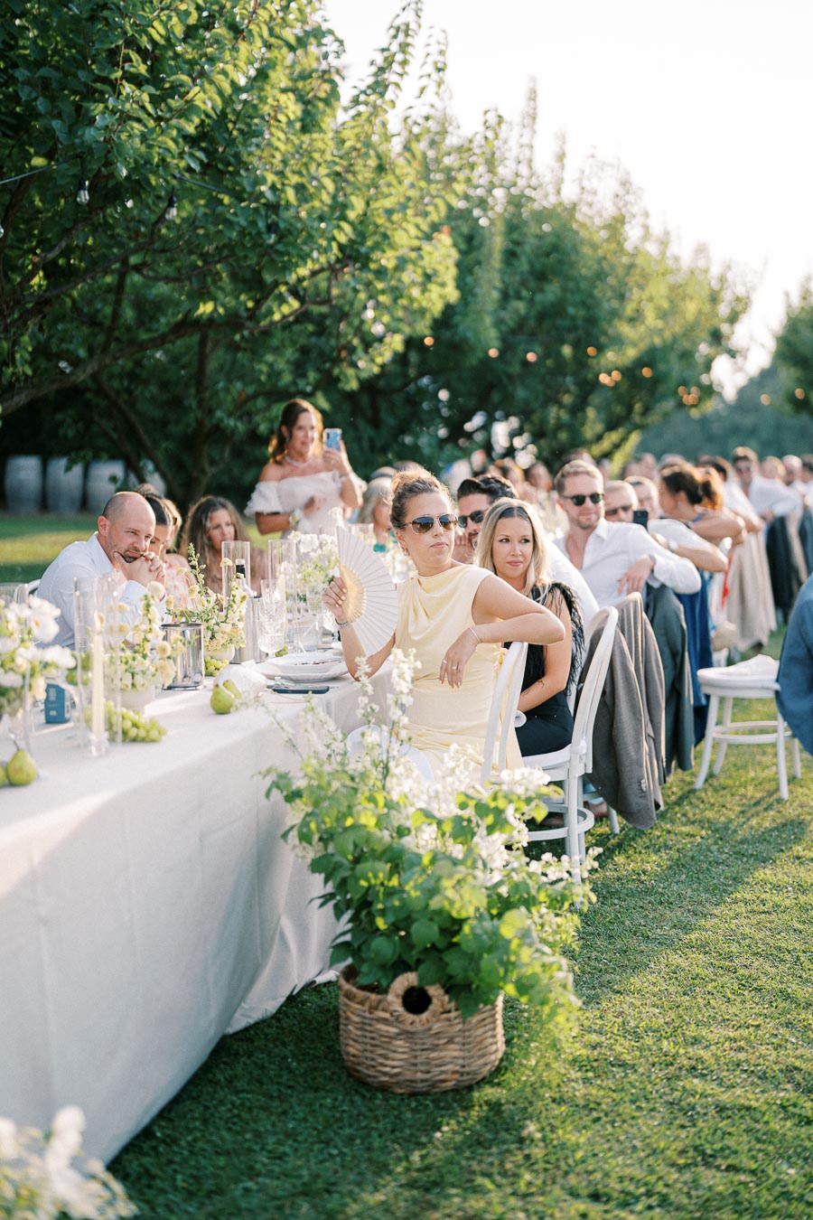 Outdoor garden party with guests seated at a long, elegantly decorated table adorned with greenery and flowers. The setting features lush trees and soft lighting, creating a serene and festive atmosphere.