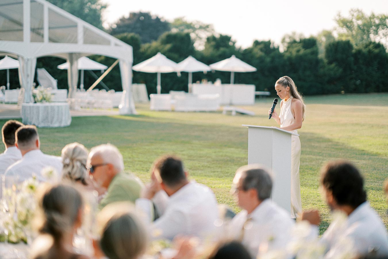 A woman in elegant attire delivers a speech with a microphone at an outdoor event, surrounded by seated guests on a sunny day, with a large white tent and umbrellas in the background.