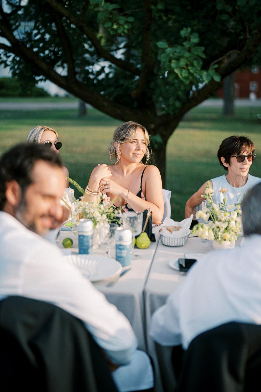 Group of people enjoying an outdoor meal at a beautifully set table under a tree, with plates, drinks, and floral arrangements on a sunny day.