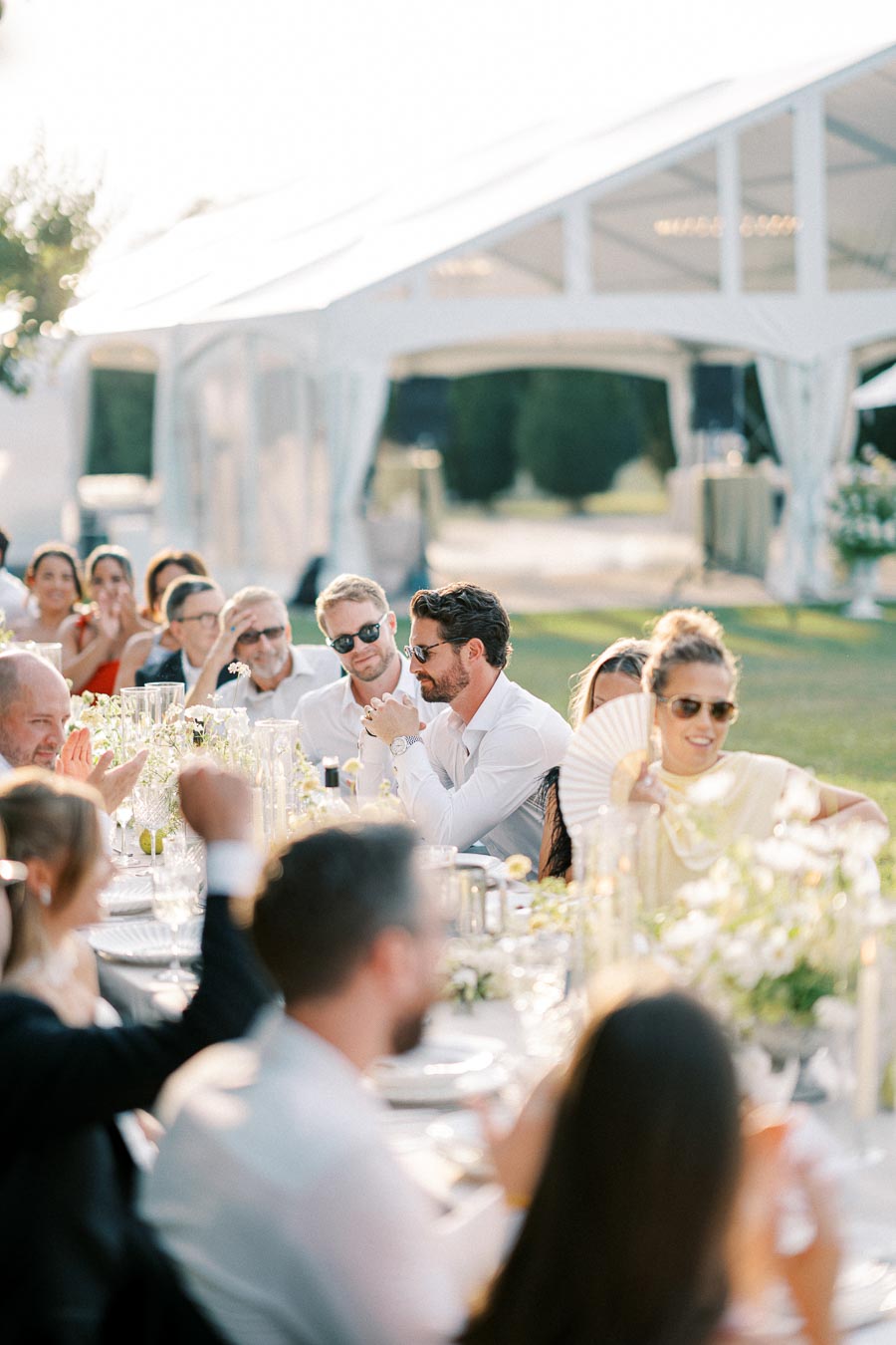 Outdoor wedding reception with elegantly dressed guests seated at a long table under a white tent. The table is beautifully adorned with flowers and candlelight, creating a festive and romantic atmosphere.