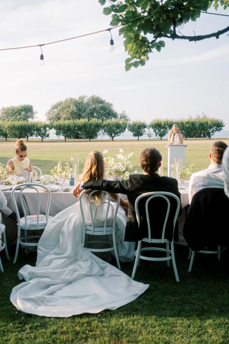 Outdoor wedding reception with bride and groom sitting at a beautifully decorated table, listening to a speech in a scenic garden setting under hanging string lights.