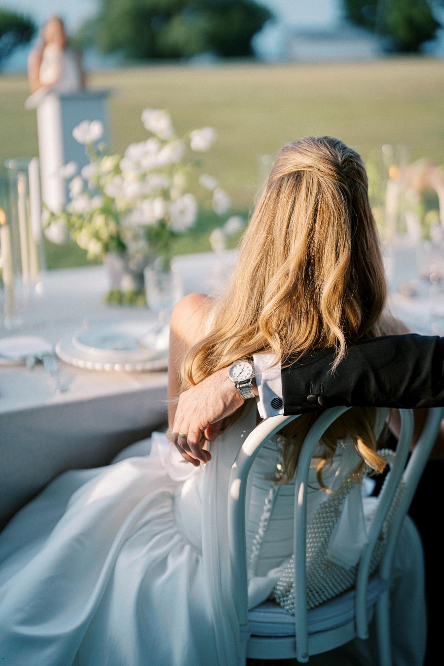 Beautiful bride with long blond hair sitting at an elegant outdoor wedding reception, adorned in a white gown, with a groom’s arm wrapped around her, showcasing a classic watch. Candlelit table setup and floral centerpieces add to the romantic atmosphere.
