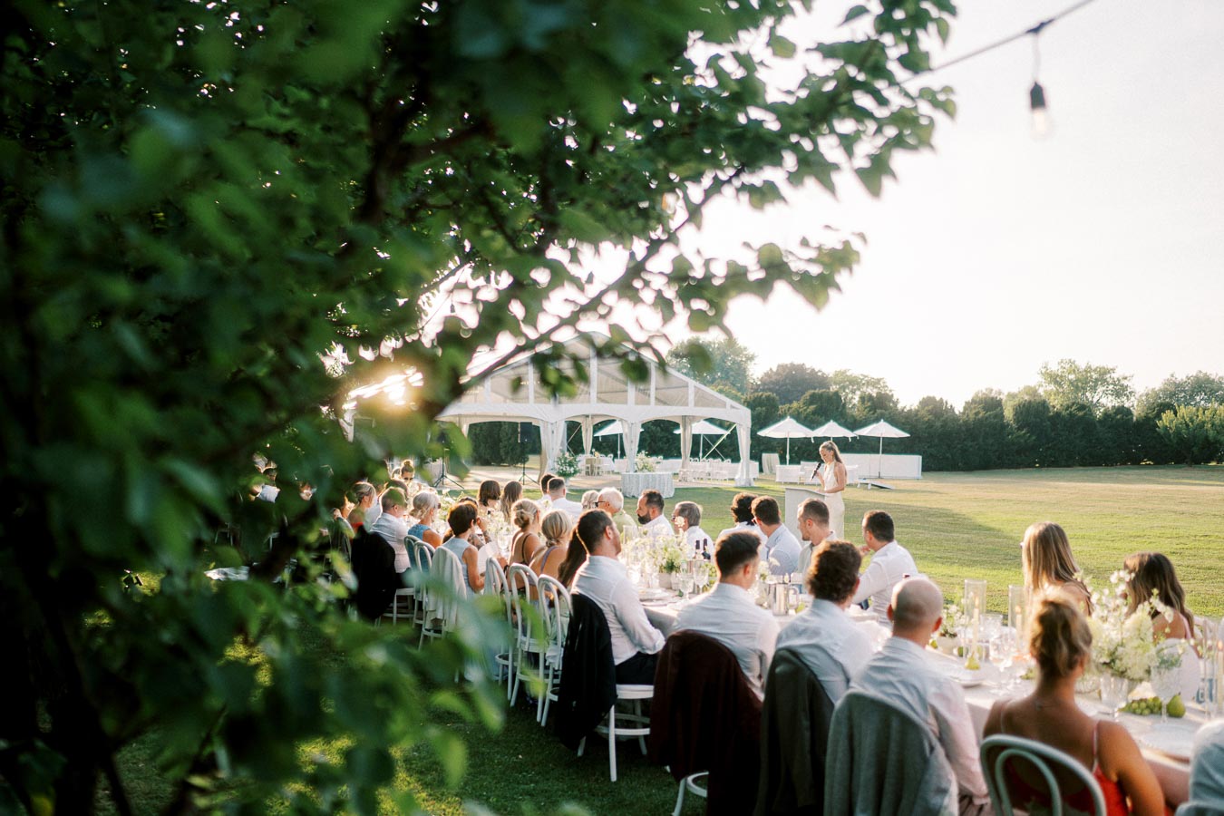 Open-air wedding reception with guests seated at long tables under trees, listening to a speech, with a white tent and greenery in the background.