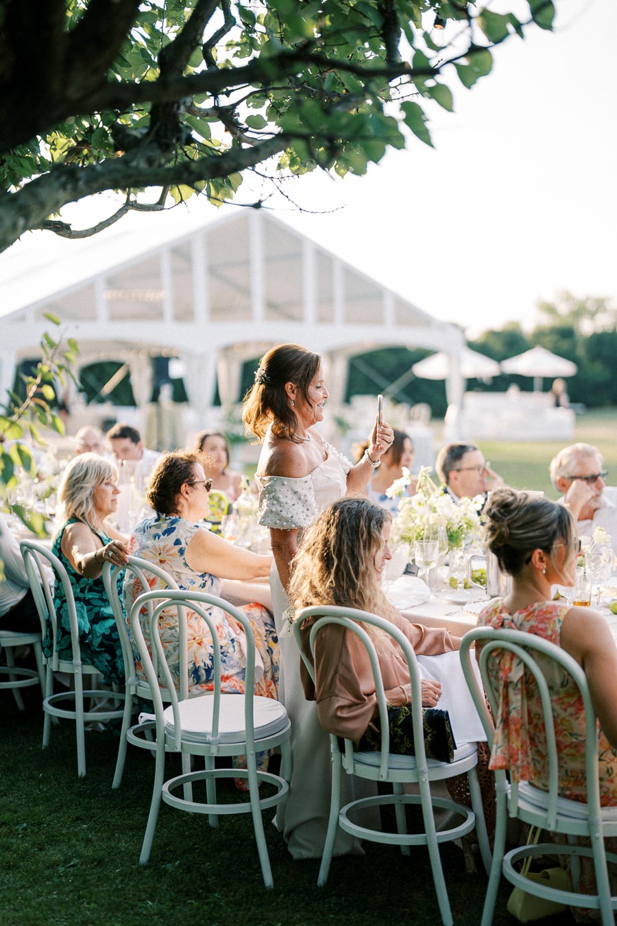 A group of elegantly dressed guests seated at a long outdoor table during a wedding reception, with a woman standing to take a photo under the shade of a tree, near a garden tent.