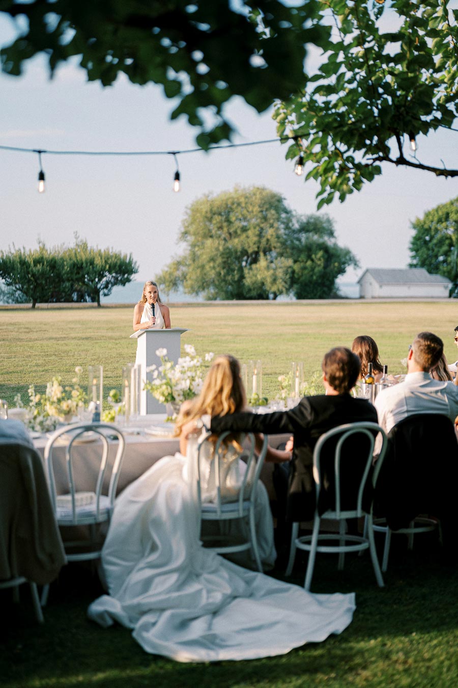 Outdoor wedding reception with a woman giving a speech at a podium; guests seated at elegantly decorated tables under string lights, enjoying a sunny day with lush trees and a scenic backdrop.