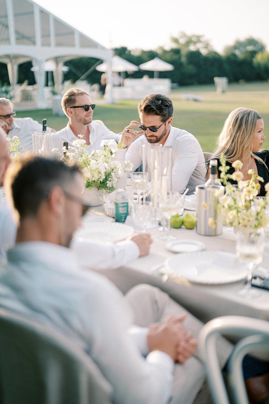 Outdoor wedding reception with guests seated at a beautifully decorated table with white flowers and elegant glassware under a clear sky.