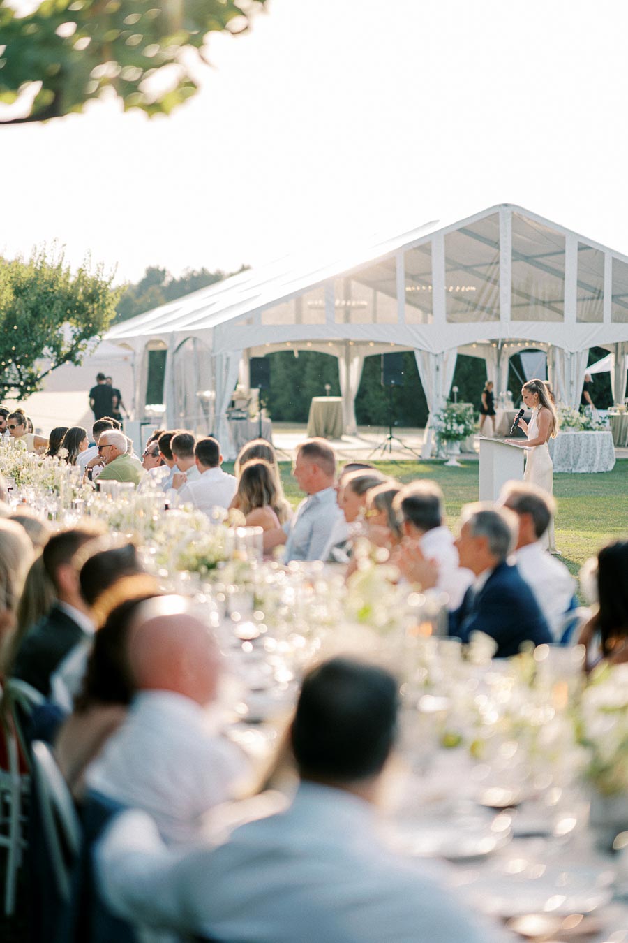 Elegant outdoor wedding reception with guests seated at a long table under a white marquee, featuring a woman giving a speech.