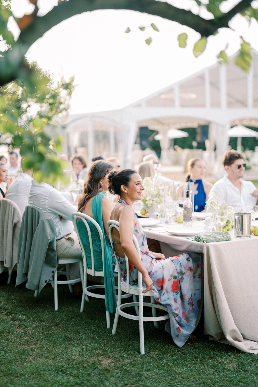 Elegant outdoor wedding reception featuring guests seated at a long table adorned with floral arrangements, enjoying a sunny afternoon in a garden setting.