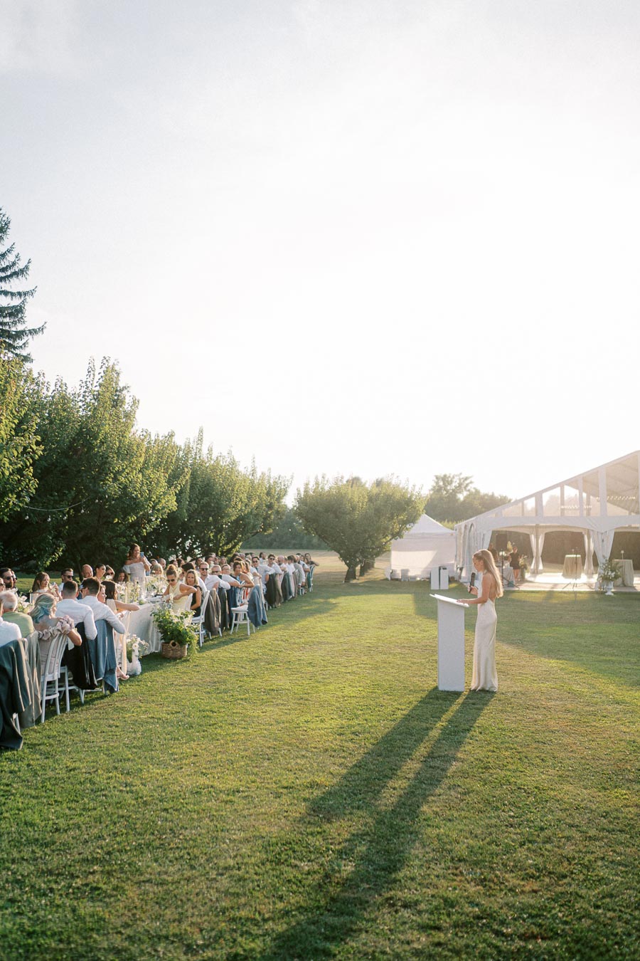 Outdoor wedding reception with long banquet table, guests seated, and a woman giving a speech under a clear sky.