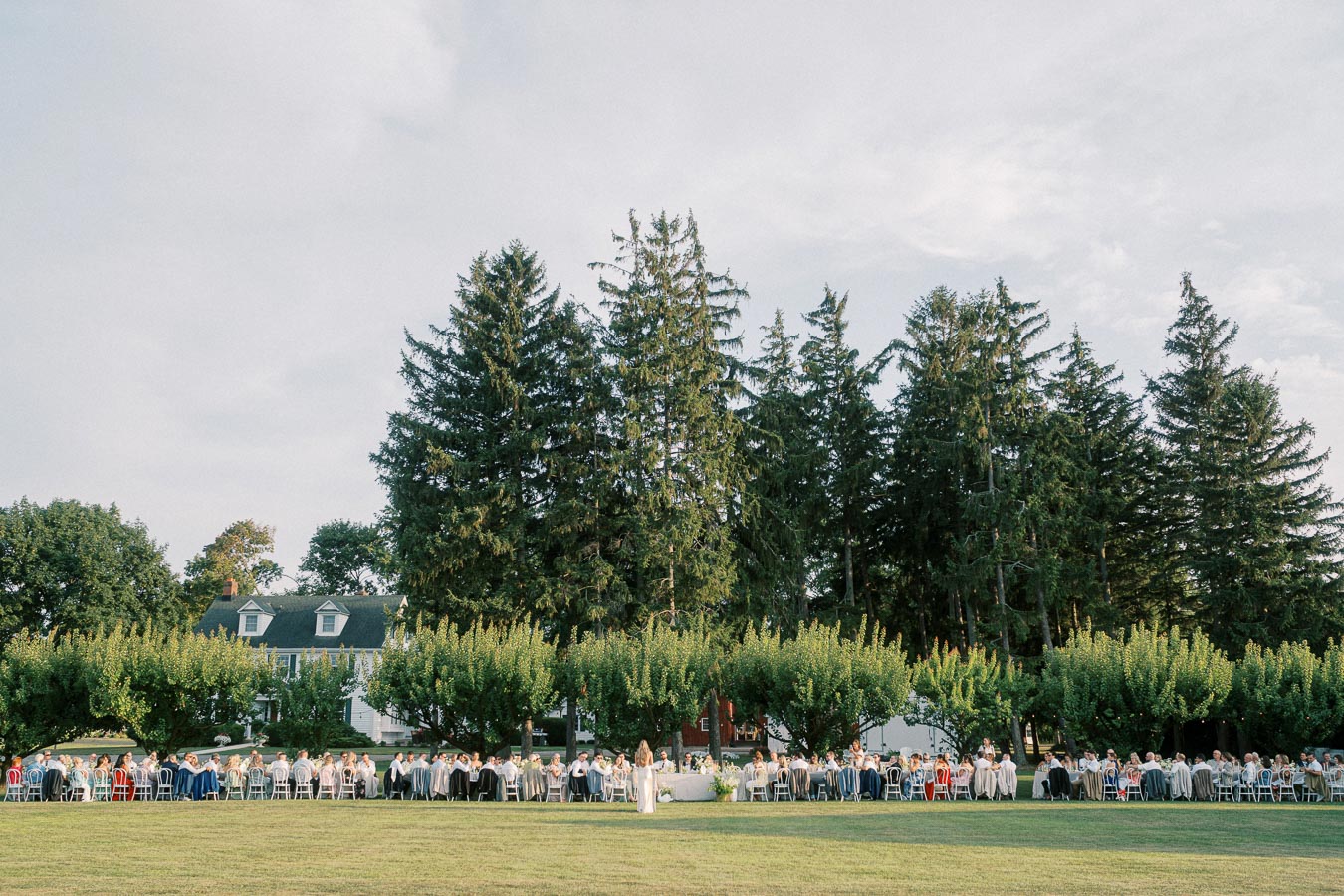 Outdoor wedding ceremony with guests seated on a grass lawn, large trees and a charming house in the background.