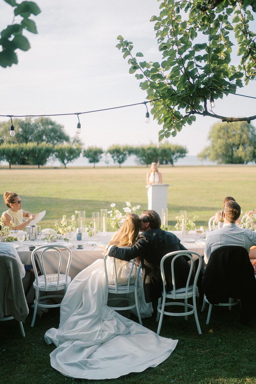 Outdoor wedding reception with a couple seated together, adorned in formal attire, under string lights. The setting features a lush green lawn, elegantly decorated tables, and a person delivering a speech in the background.