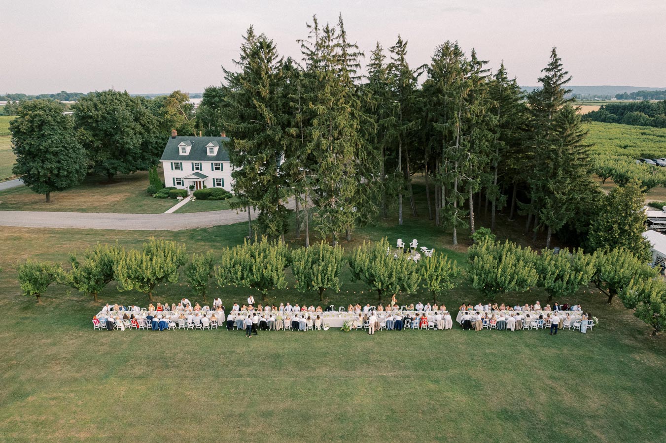 Aerial view of an outdoor event with long tables and guests seated in a scenic countryside setting, surrounded by trees and a large house in the background.